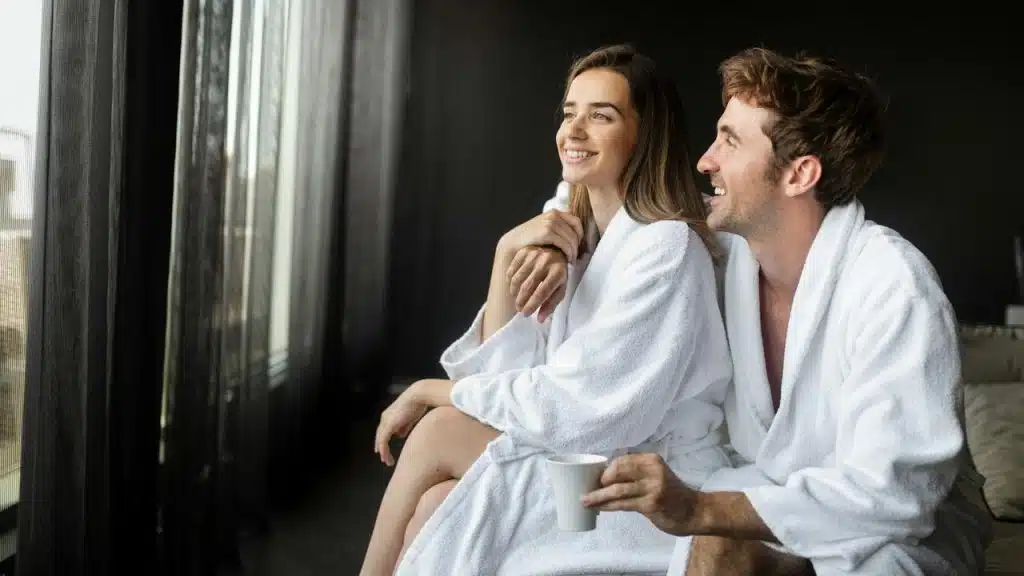 A couple in white bathrobes sitting by a window, smiling and enjoying a drink.