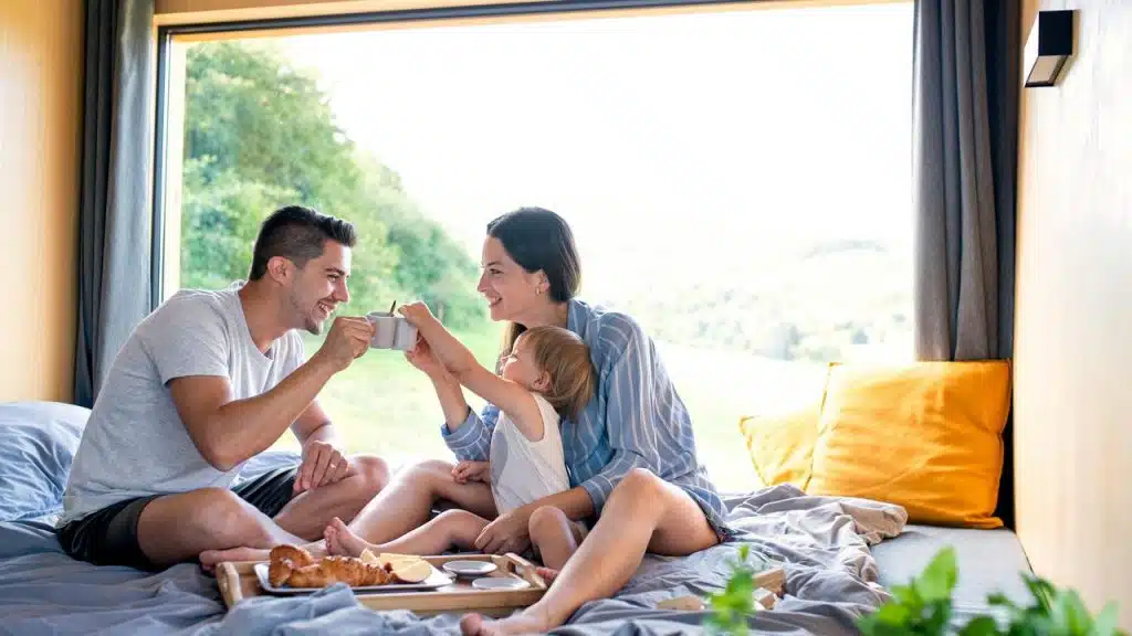 A family sitting on a bed by a large window, sharing breakfast and smiling together.