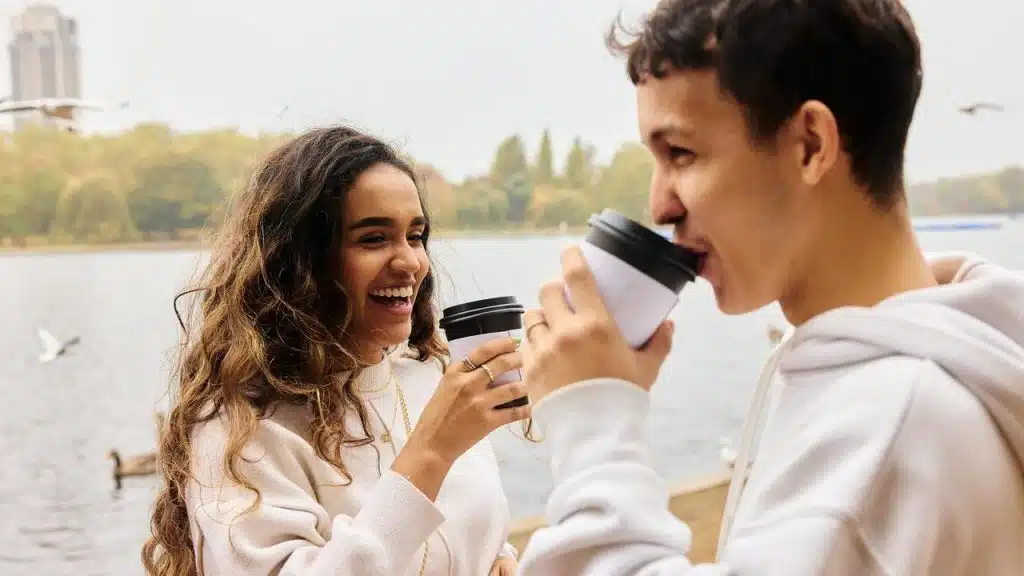 A couple smiling and enjoying coffee together by a lake.