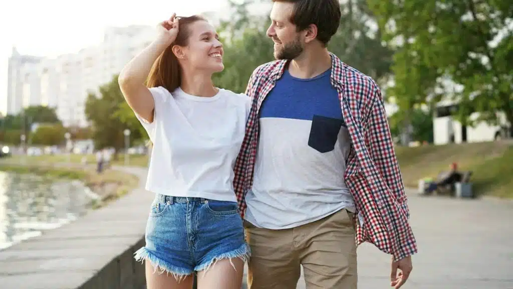 A couple walking together by the water, smiling and looking at each other.