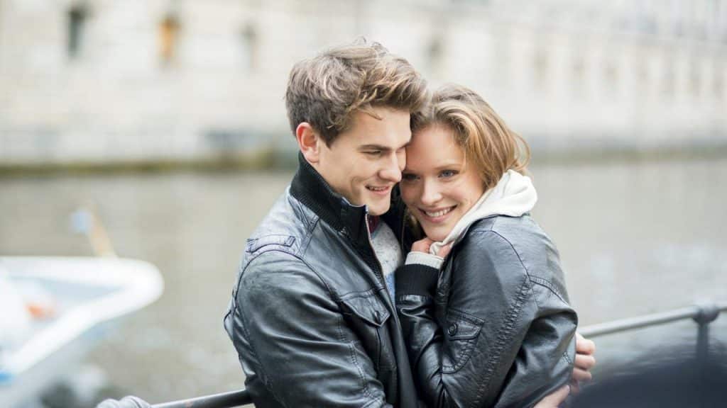 A couple embracing and smiling by the water on a cloudy day.
