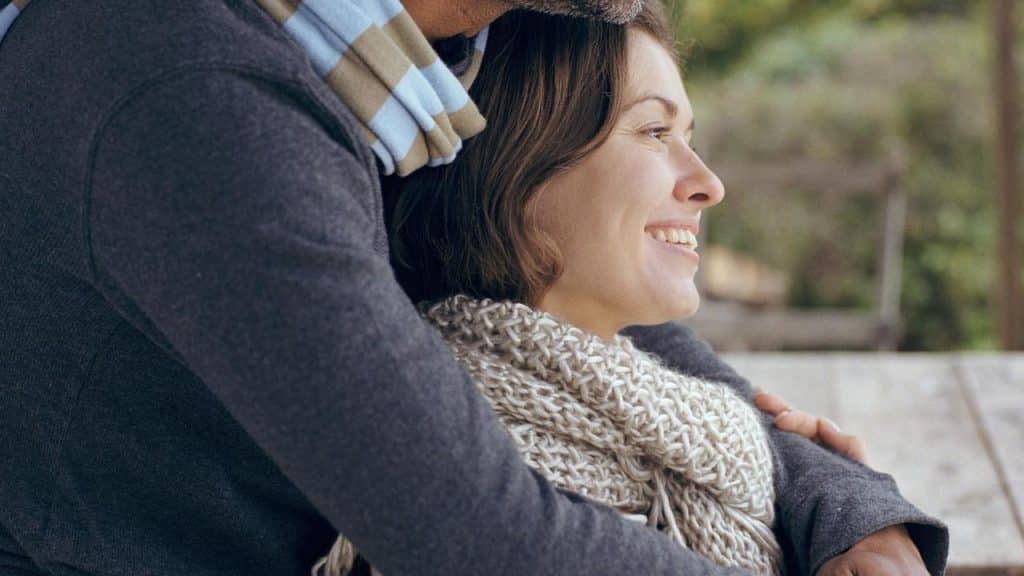 A woman smiling while being warmly embraced by a man outdoors.