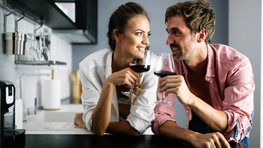A couple in a kitchen smiling at each other while holding glasses of red wine.