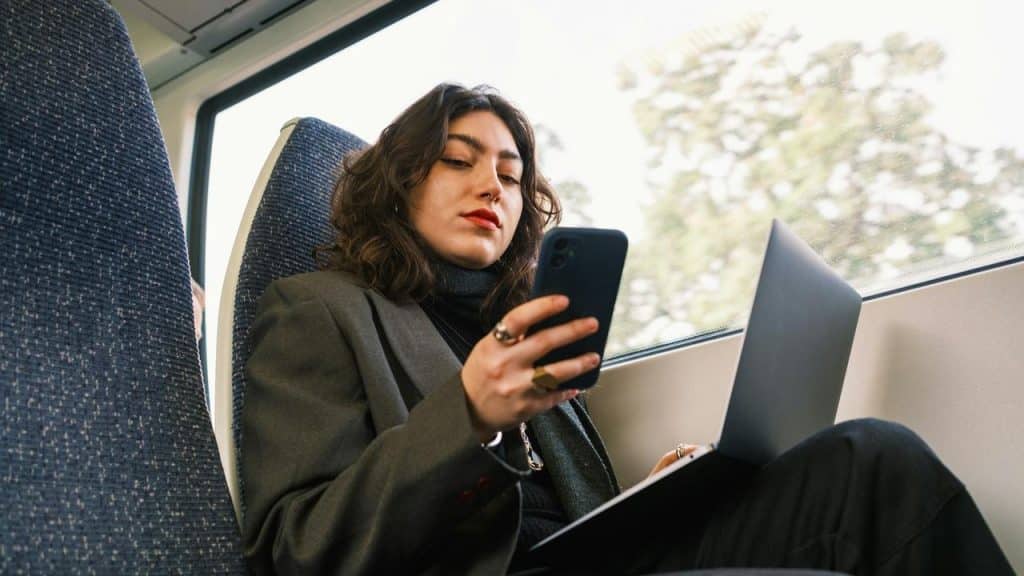 A woman sitting on a train seat using her phone while holding a laptop on her lap.