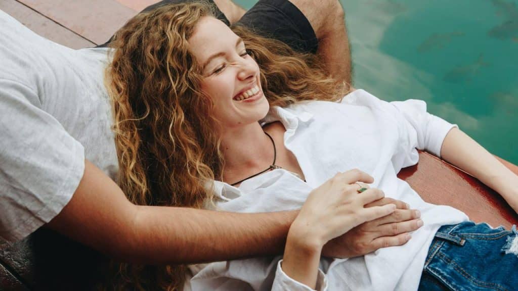 A smiling woman lying on a dock with someone’s arm around her.
