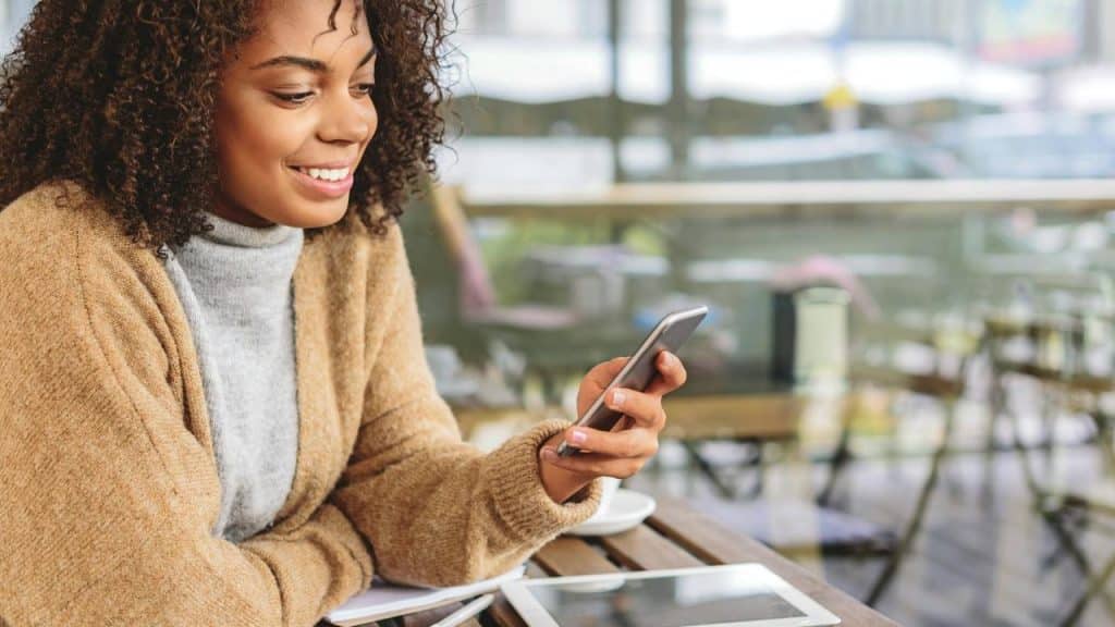 A smiling woman sitting at an outdoor café looking at her phone.