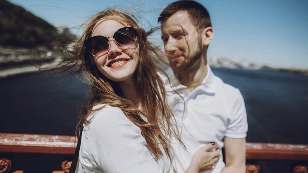 A smiling woman wearing sunglasses standing next to a man near the water on a windy day.