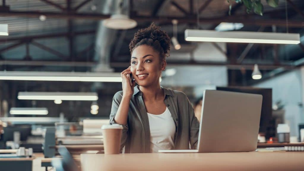 A smiling woman sitting at a desk in an office, talking on her phone.