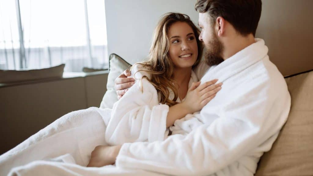 A couple wearing white bathrobes sitting close together and smiling at each other.