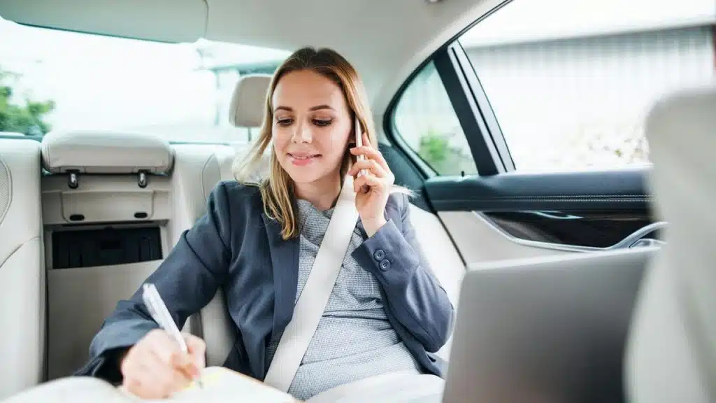 A woman sitting in the back seat of a car talking on the phone while writing in a notebook.