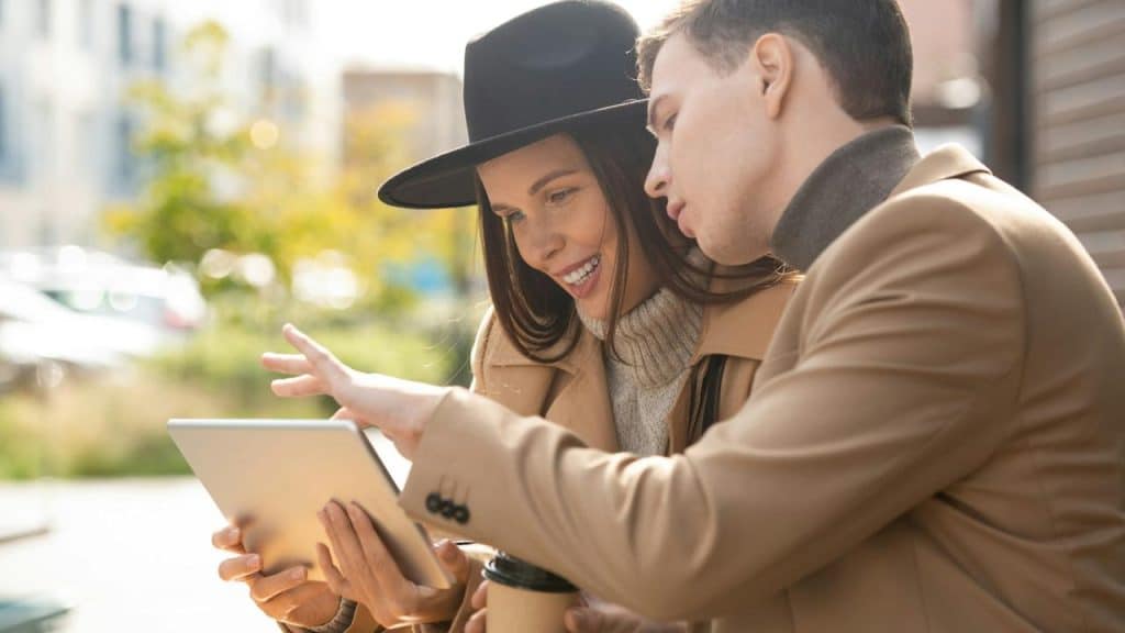 A smiling couple sitting outdoors looking at a tablet together.