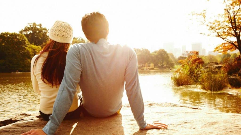 A couple sitting by a lake facing the sunlight.