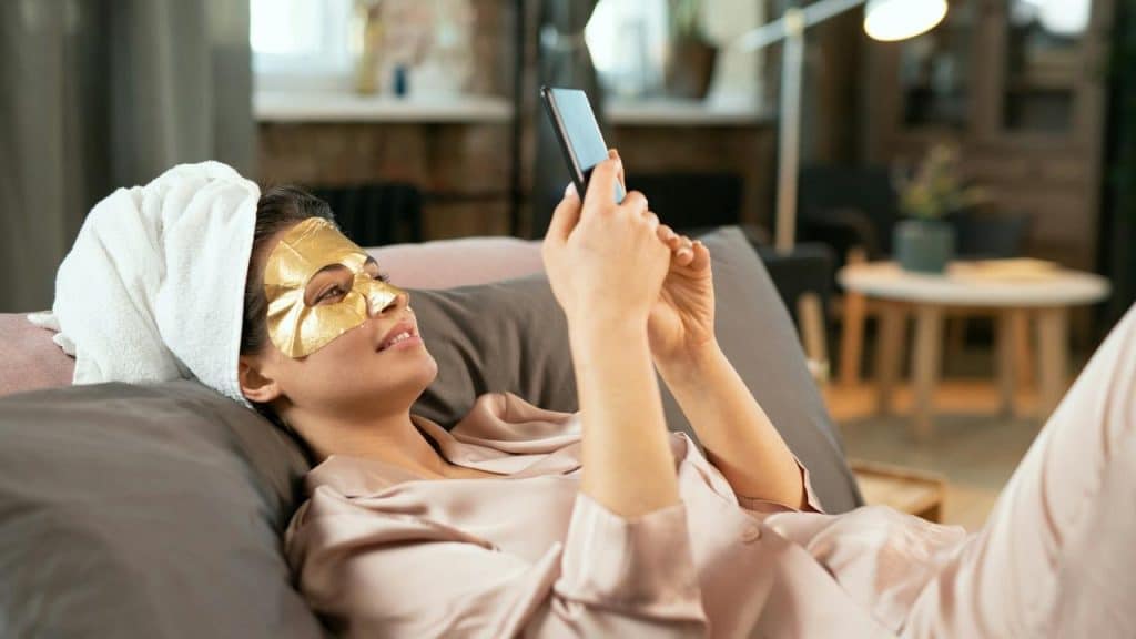 A woman lying on a couch wearing a gold face mask and using her phone.