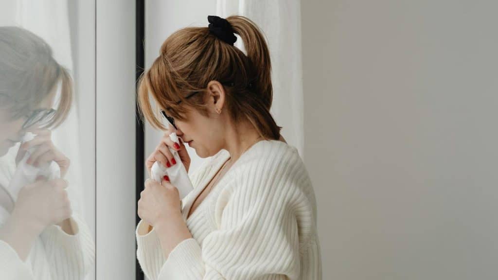 A woman holding a tissue and appearing emotional by a window.