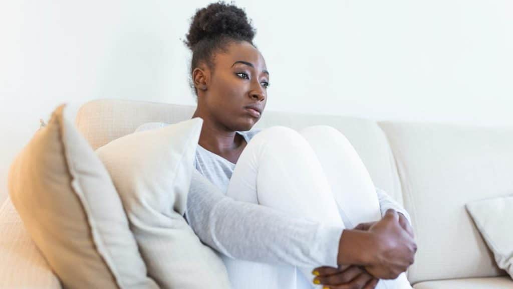 A woman sitting on a couch hugging her knees, looking sad.