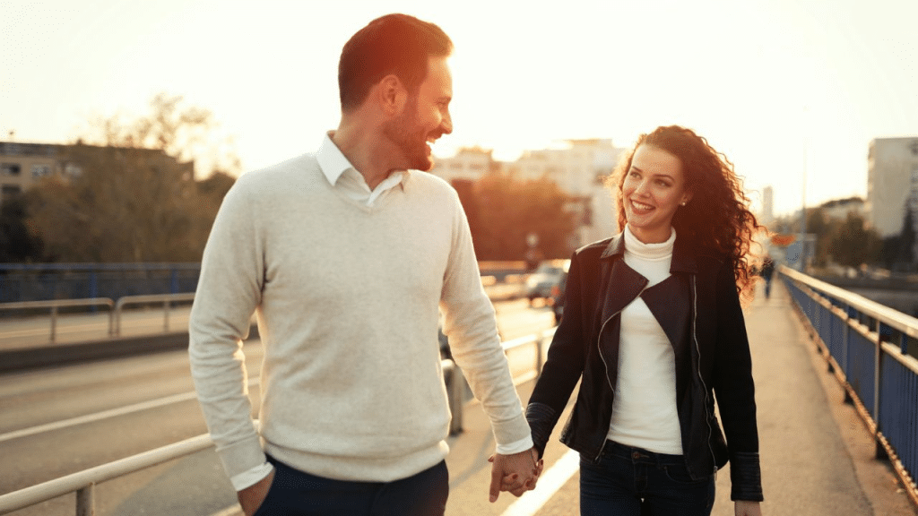 A happy couple holding hands and smiling at each other while walking down a bridge at sunset.