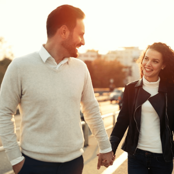 A happy couple holding hands and smiling at each other while walking down a bridge at sunset.