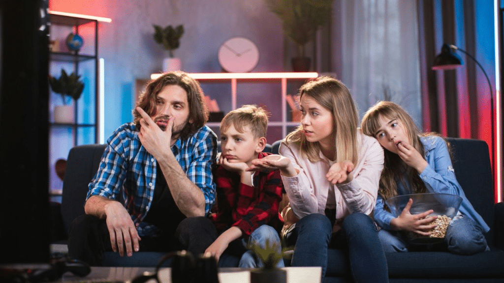 A family of four sits on a couch, seemingly confused or displeased while watching something.