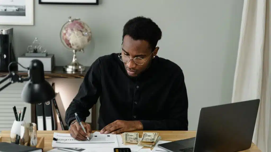 A man working at a desk with papers, cash, and a laptop.