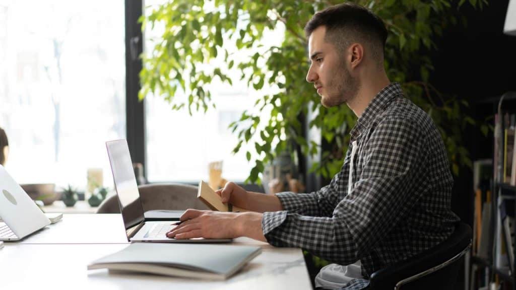 A man sitting at a desk using a laptop and holding a notebook.
