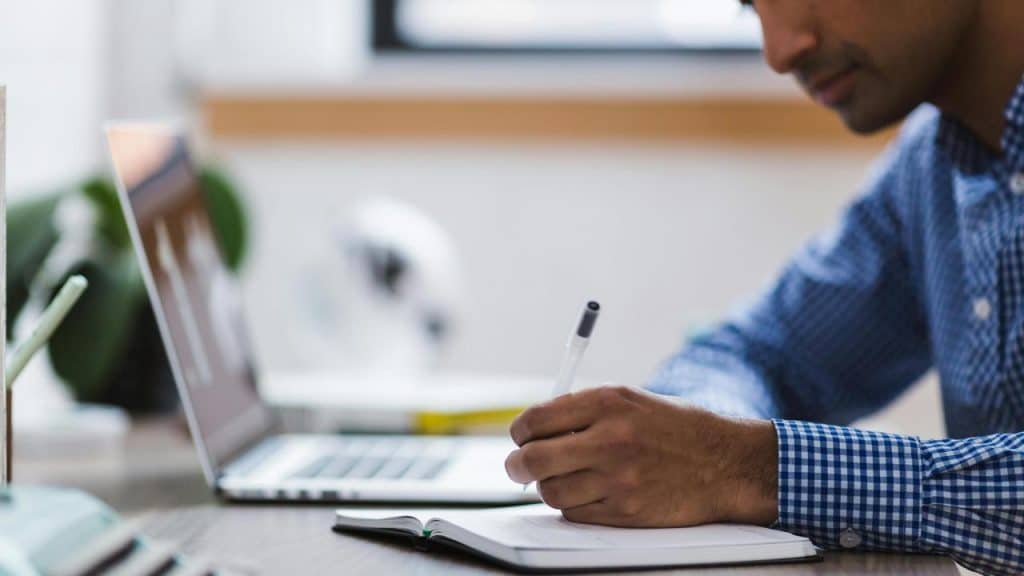 A man writing in a notebook while working on a laptop.