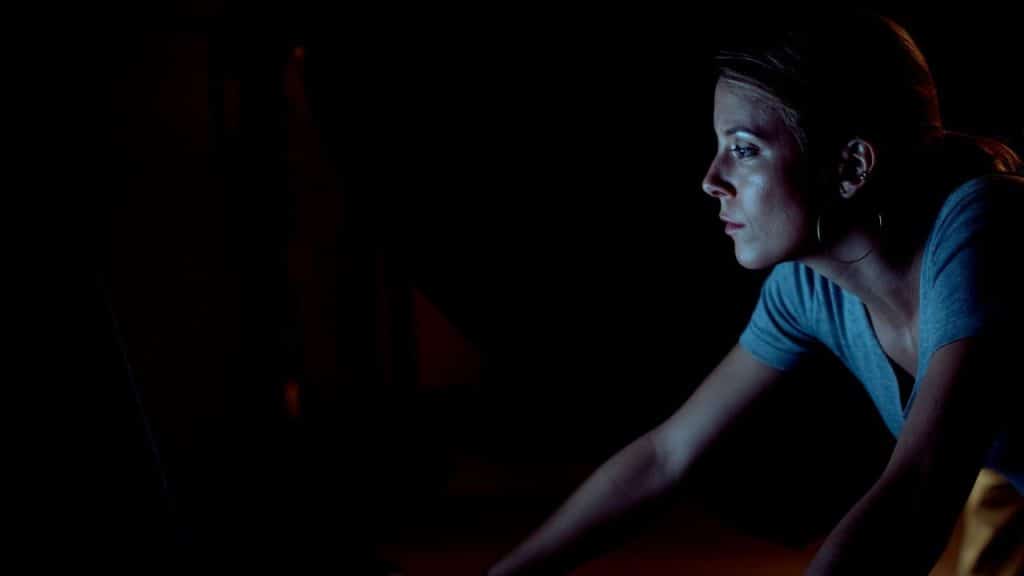 Woman's face lit by a screen in a dark room, looking intently.