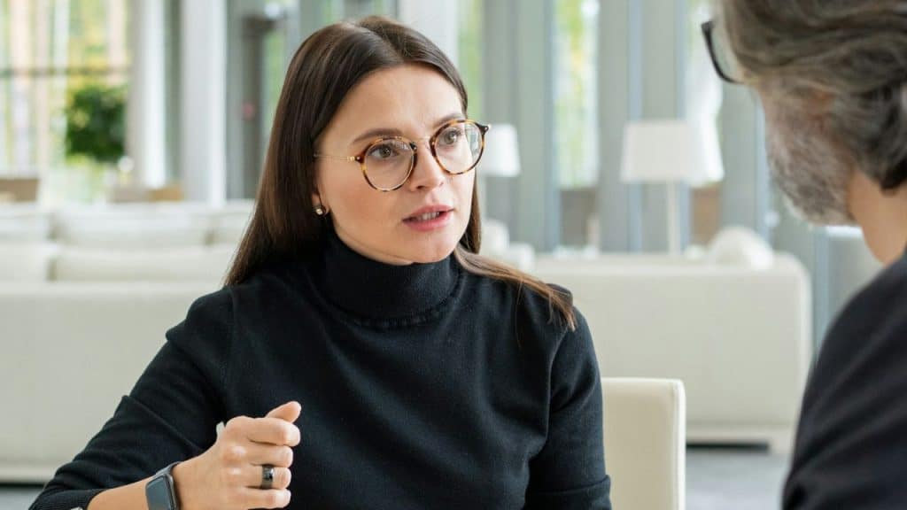 Woman in glasses and a black turtleneck speaking to an unseen person indoors.