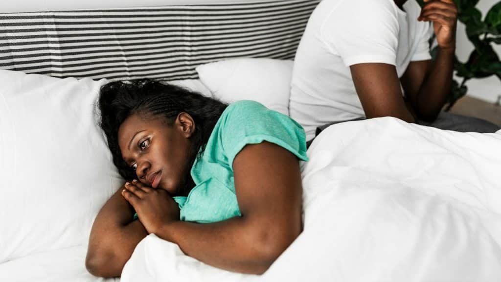A distressed Black woman in bed, facing away from a Black man sitting behind her.