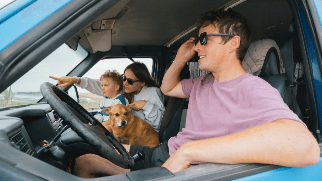A family, including a man, woman, child, and dog, sits in a blue car. The woman is pointing something out the window.