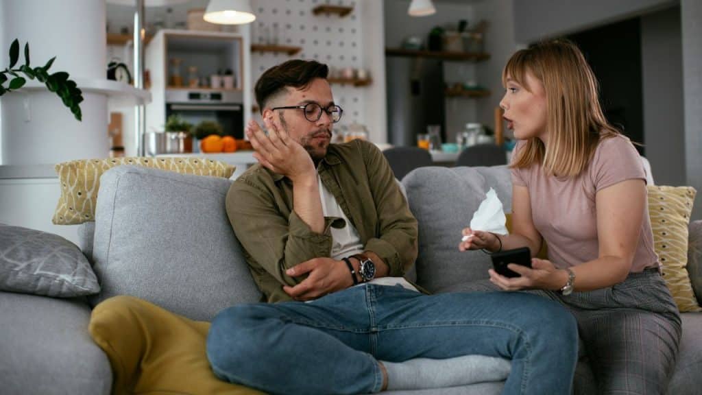 A man and a woman talking on a couch; the woman holds a phone and tissue.