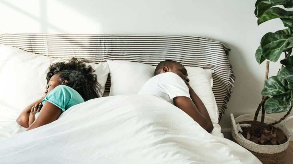 A Black man and woman lying in bed, facing away from each other under white covers.