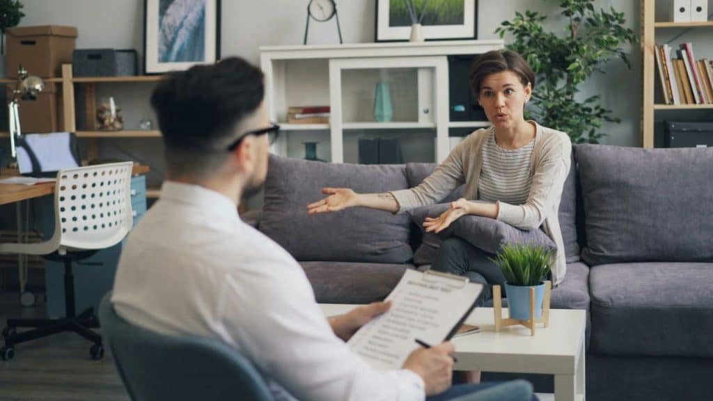 A woman talks animatedly to a man holding a clipboard, possibly in therapy.