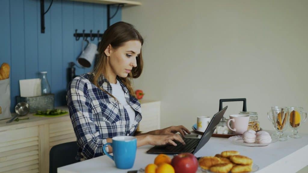 A woman in a plaid shirt is focused on typing on a laptop in a kitchen.