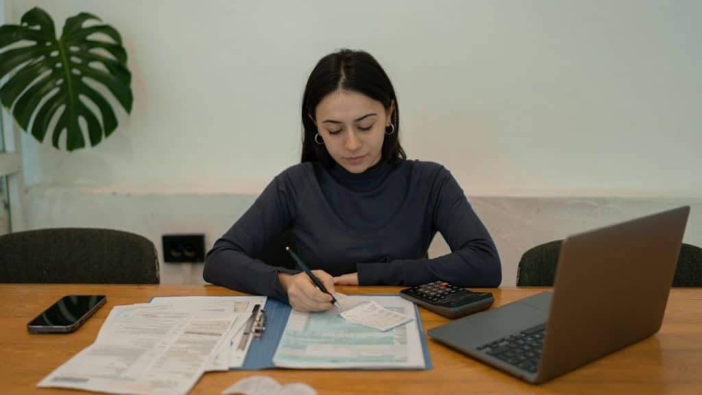 A young woman with dark hair is writing on a form with a laptop and calculator nearby.