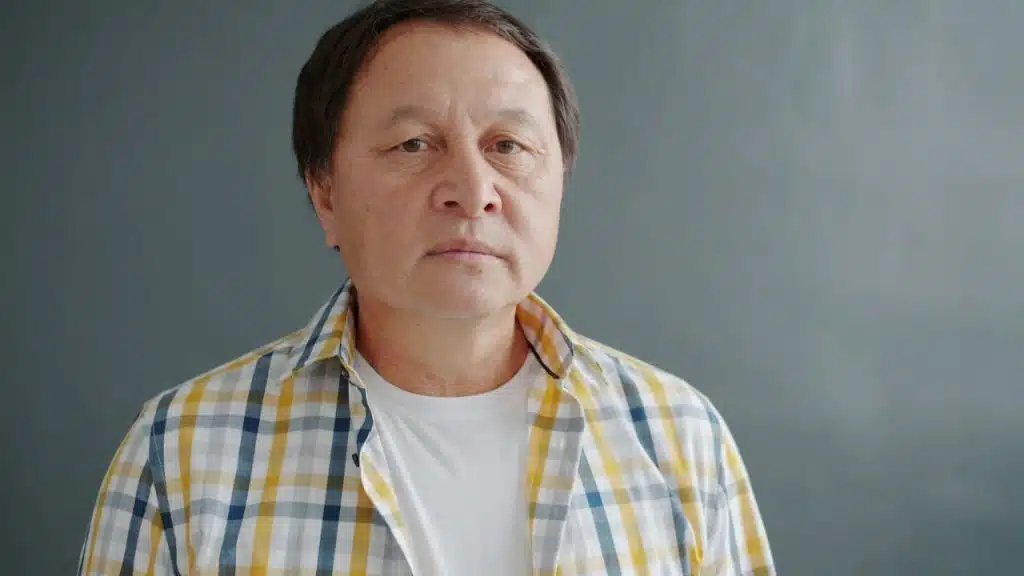 A middle-aged man in a plaid shirt and white t-shirt looks directly at the camera.