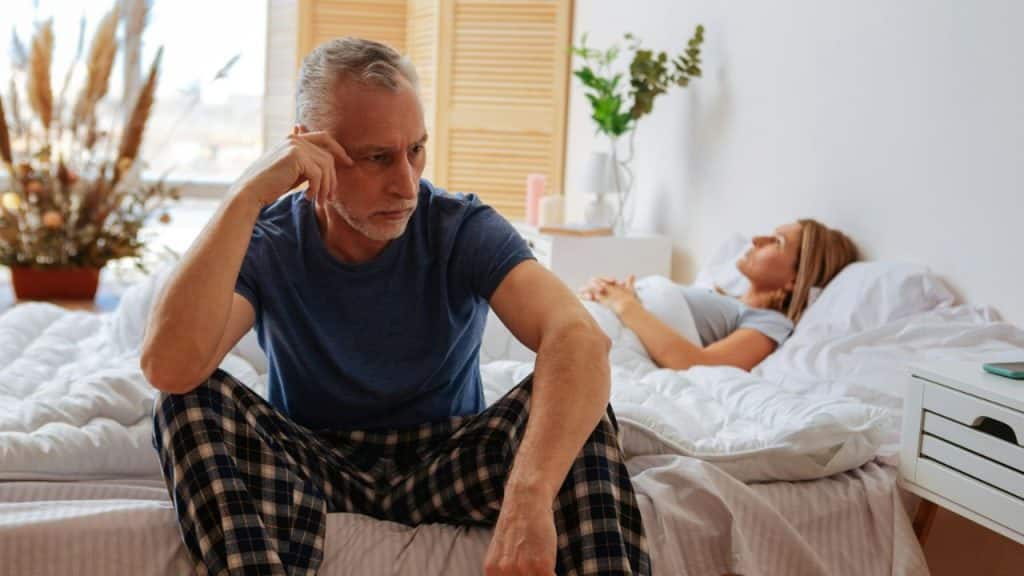 A worried older man sits on a bed with a woman lying down behind him.