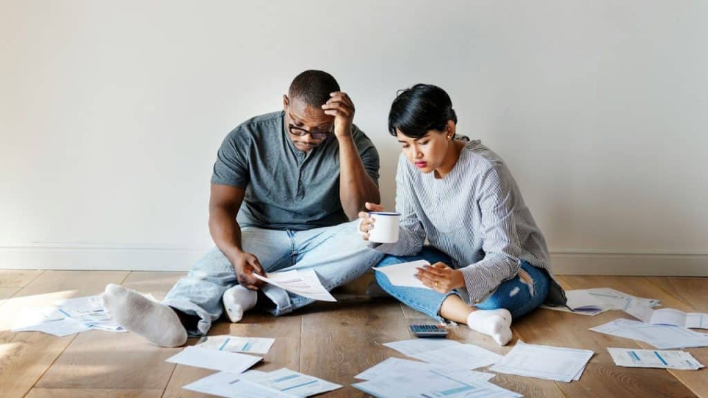 A Black man and an Asian woman sitting on the floor looking stressed at financial documents.