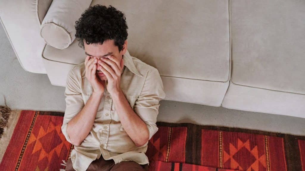 A man with curly hair sits on the floor, covering his face with his hands.
