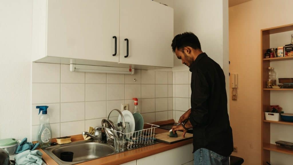 A man in a black shirt is cutting vegetables next to a sink full of dishes.