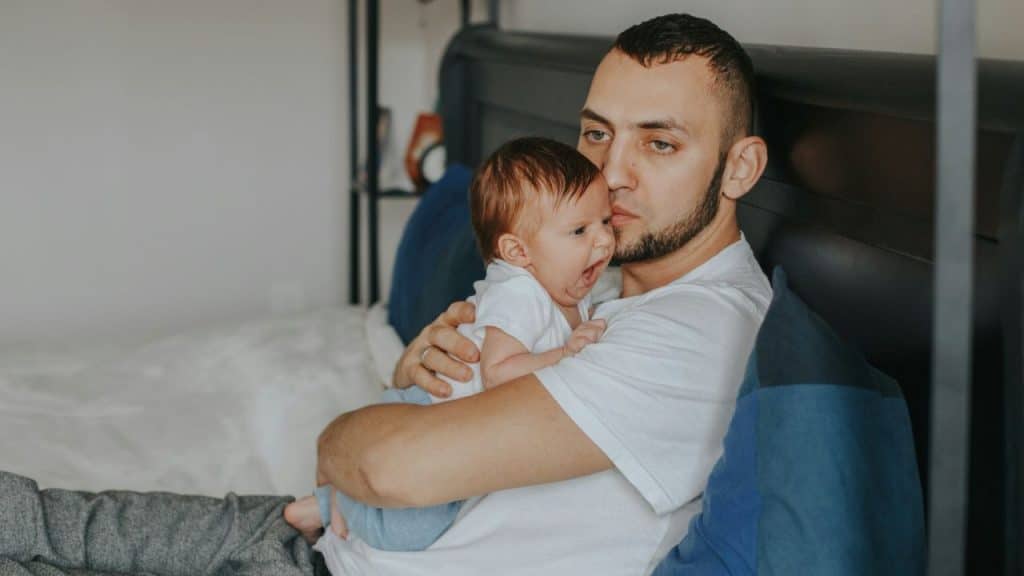 A man with a beard holds a crying infant while sitting on a bed.