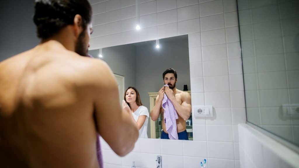 A shirtless man looks in a mirror with a woman standing behind him in the reflection.