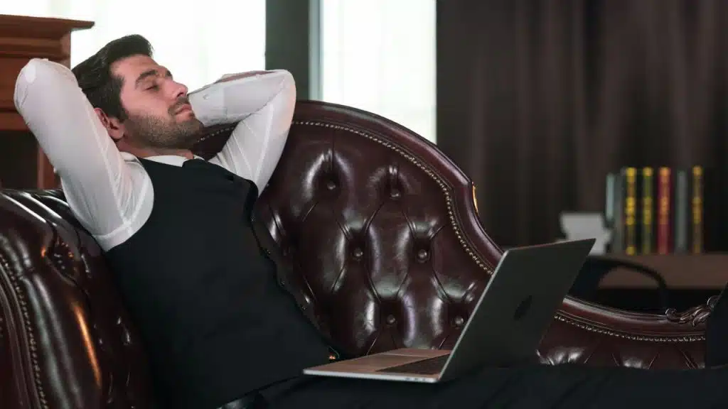 A man in a vest and white shirt reclines on a leather couch with a laptop on his legs.