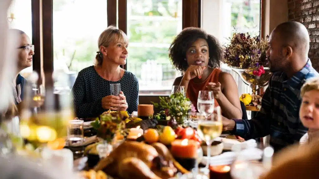 A diverse group of adults and a child are talking around a dinner table with food.