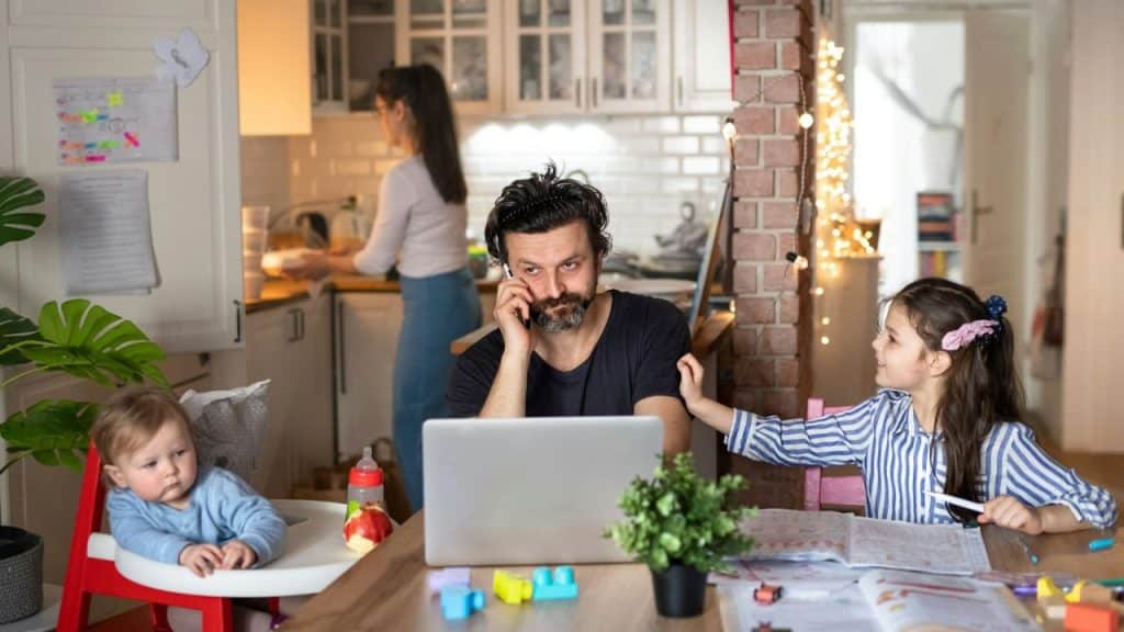 A busy father works on a laptop at a table with two children while a woman works in the background.