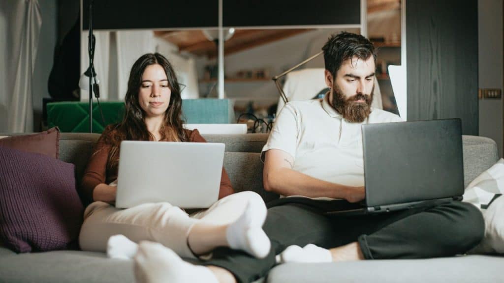 A woman and a bearded man sit on a couch using separate laptops.
