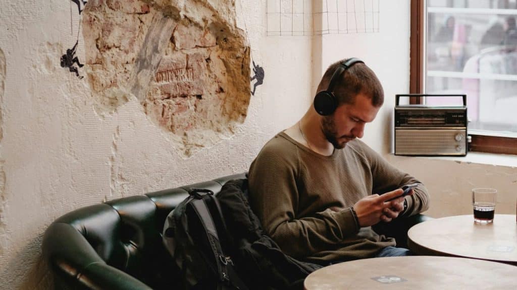 A man wearing headphones looks at his phone while sitting on a dark green couch.