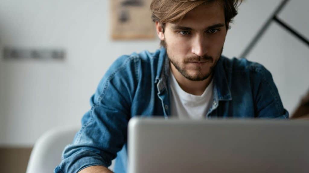 A focused, bearded man in a denim shirt looks intently at a laptop screen.