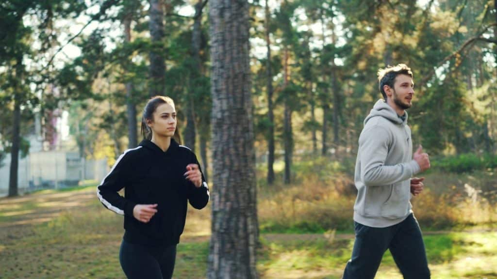 A man and a woman are jogging side-by-side on a sunny, wooded path.