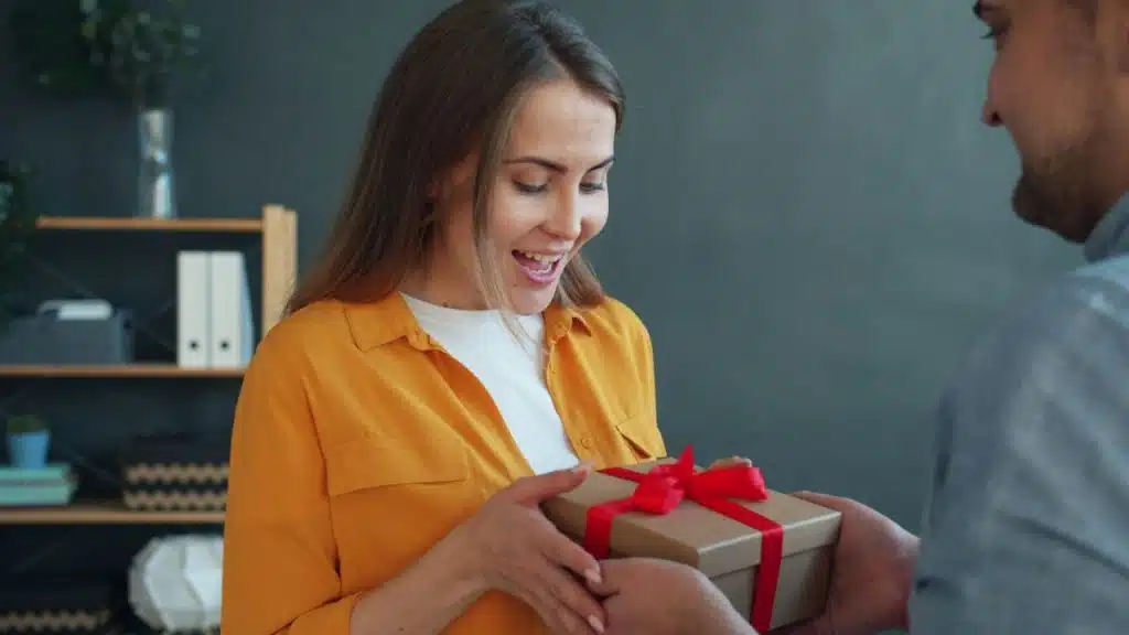 A surprised woman in an orange shirt is happily receiving a gift box from a man.