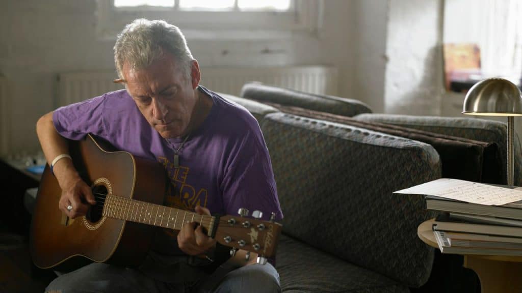 A middle-aged man in a purple shirt sits on a couch and plays an acoustic guitar.